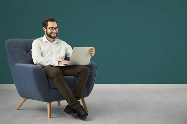 Handsome man with laptop sitting in comfortable armchair against color wall min